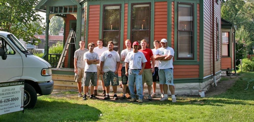 A group of paint and dirt covered individuals gather outside of a Historical Home after working on a restoring the exterior.